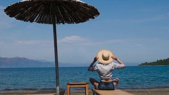 穿着衬衫和帽子的女孩坐在沙滩上的日光浴床上撑着一把伞