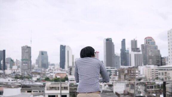 年轻的亚洲商人站在办公楼屋顶的露台上看着背后的城市景色城市生活方式