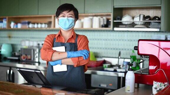 一位亚洲华人小企业主戴着口罩和防护手套在厨房柜台前微笑着看着镜头把甜点递给她的顾客