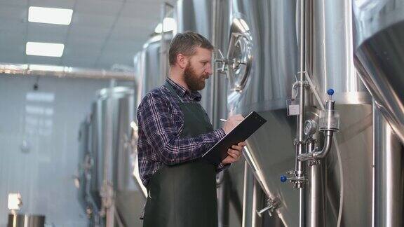 一个留着胡子的男性酿酒师站在啤酒罐旁边记录仪器读数啤酒酿造工艺