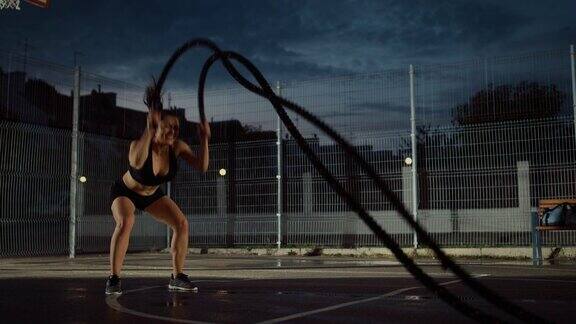 美丽的精力充沛的健身女孩做运动与战斗绳她正在一个带围栏的室外篮球场里锻炼雨后住宅区的晚间录像