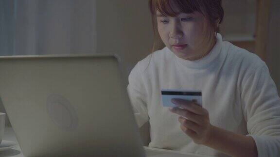 美丽的亚洲女人使用笔记本电脑购买网上购物通过信用卡而穿着随意坐在客厅的桌子上晚上在家里生活方式女性在家办公