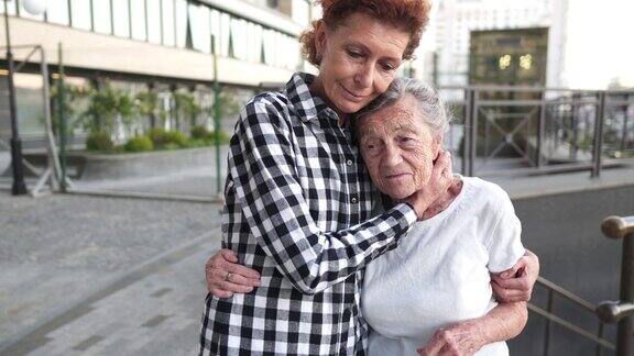 奉献精神在城市中漫步的完美伴侣照顾老人的志愿者助理高层妇女与女儿拥抱和牵手在多层建筑的院子里