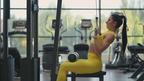 一名西班牙妇女坐在健身房的模拟器上用重量拉动一根金属绳带动背部肌肉黑发女人打开模拟器进行背部肌肉模拟训练