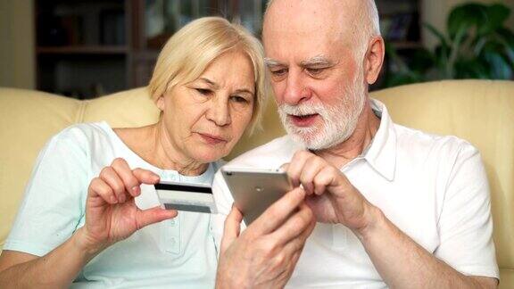 一对兴高采烈的好看的老夫妇坐在沙发上在智能手机上用信用卡在线购物