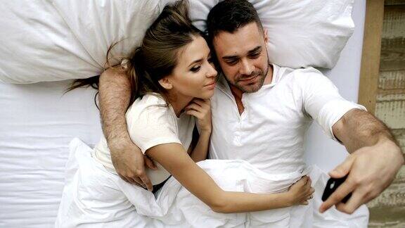 年轻美丽的情侣用智能手机自拍早上躺在床上接吻