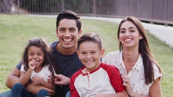快乐的年轻西班牙家庭坐在公园的草地上互相挠痒痒对着镜头微笑