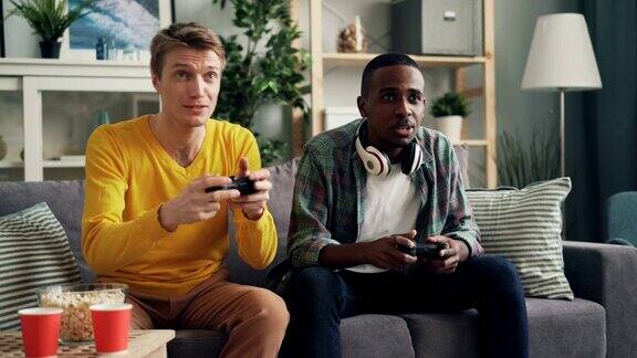 两个不同种族的朋友坐在家里的沙发上玩电子游戏