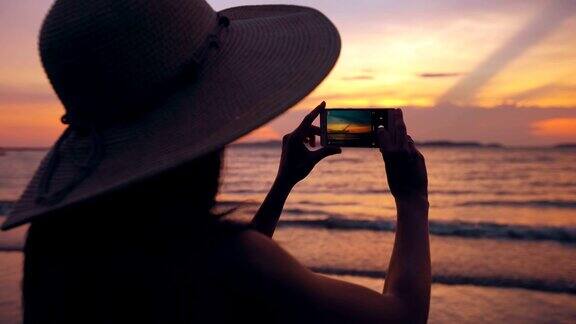 年轻女游客的剪影戴着帽子拍照与手机在日落在海洋海滩