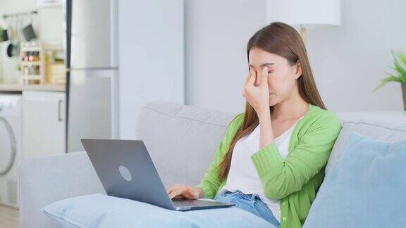精疲力竭的亚洲女商人在使用笔记本电脑时伤了眼睛年轻的女白领坐在沙发上感到视觉疲劳和眼睛疲劳过度在家按摩干燥易怒的眼睛