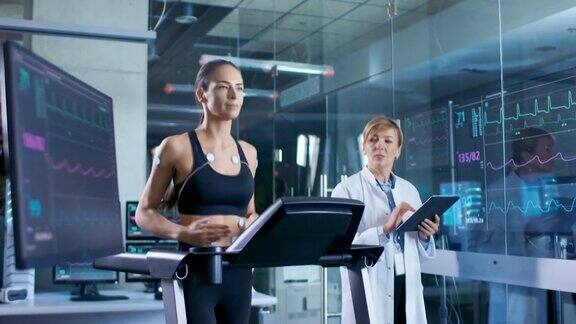 美丽的女人运动员在跑步机上跑步与电极连接到她的身体女医生使用平板电脑和控制心电图数据显示在实验室监视器