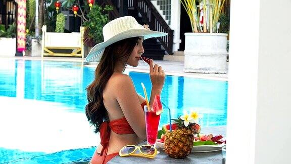 夏天性感的亚洲女人在橙色比基尼吃玫瑰苹果而放松在游泳池