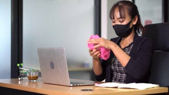 亚洲办公室女性戴口罩使用喷雾消毒酒精消毒和擦拭工作场所的笔记本电脑清洁工作场所的办公桌预防冠状病毒COVID-19