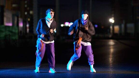 两个年轻美丽的女人跳嘻哈的二重奏自由式在夜晚的城市的街道上青年的生活方式和情感观念