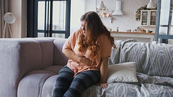 年轻的超重妇女忍受着腹痛经期痉挛坐在家里的沙发上