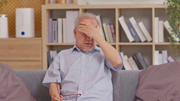 亚洲老年老资格男子头痛和偏头痛老人身患病痛独自坐在家里的沙发上深思熟虑、压力过大和孤独会导致抑郁和不良的心理健康