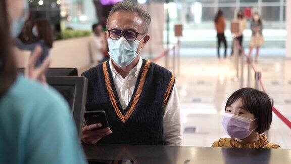 亚洲华人爷爷孙女戴着口罩在电影院的售票处用手机app购买电影票