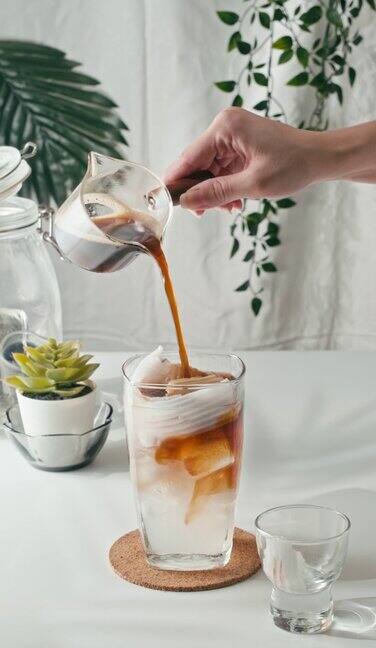 垂直4k将冲泡的咖啡倒入装满冰块的玻璃杯中木桌上有饼干用慢动作制作冷咖啡饮料喝椰汁混合浓缩咖啡从杯子短