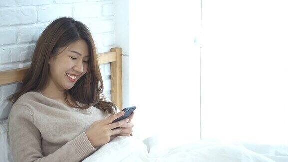 快乐的亚洲女性早上在床上使用智能手机一名亚洲女性在床上用智能手机查看社交应用微笑的女人在家里用手机上网手机成瘾