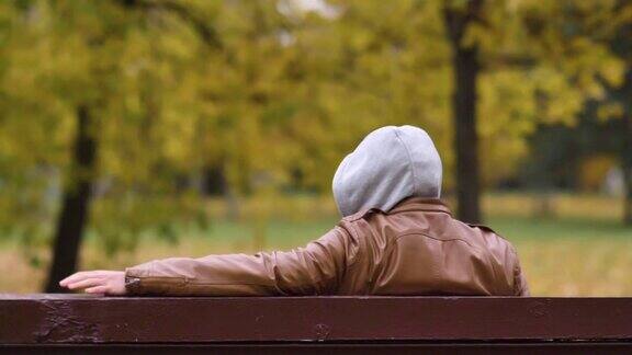 一个戴着兜帽的不知名的危险人物坐在秋天公园的长椅上看着