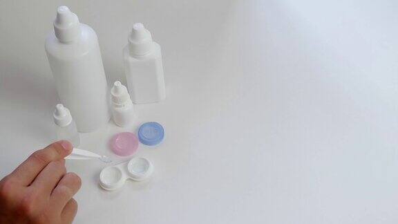 一只拿着镊子的女性手将隐形眼镜放在一个塑料容器中放置在白色的背景上还有小瓶和装有眼药水的瓶子每日镜片护理