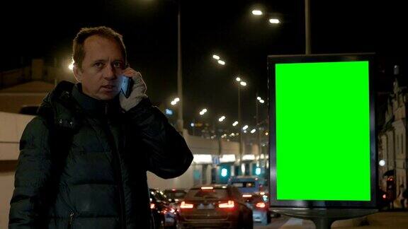 一个男人正在城市里讲电话智能手机是一种通讯工具在背景的衬托下有一个带绿色屏风的广告牌