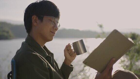 快乐的男性远足者在湖边边喝咖啡边看书