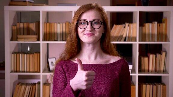 一个年轻的红发迷人的白人女学生在眼镜显示一个拇指向上微笑看在大学图书馆的相机特写肖像