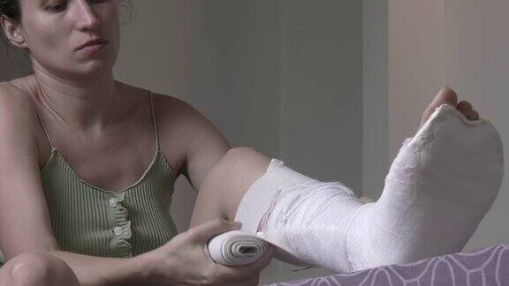 一个女人坐在床上给断了的腿绑上弹性绷带腿上打了石膏腿部骨折后的家庭康复高质量的全高清镜头