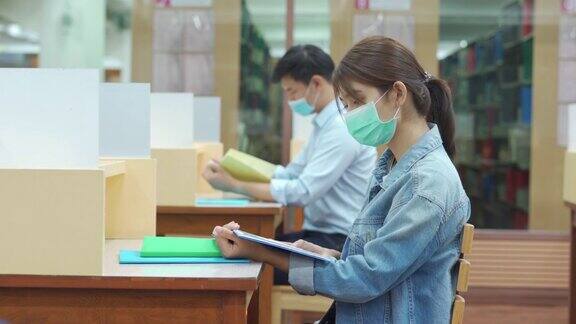两名亚洲大学生戴口罩坐在图书馆社交距离6英尺的地方以避免新冠病毒在大学内传播