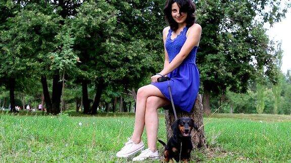 穿着蓝色裙子的女孩和一只腊肠狗坐在公园的树桩上