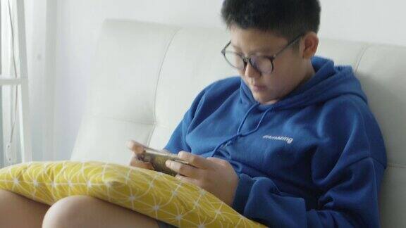 亚洲男孩戴着眼镜坐在家里的沙发上玩智能手机网络游戏生活方式概念