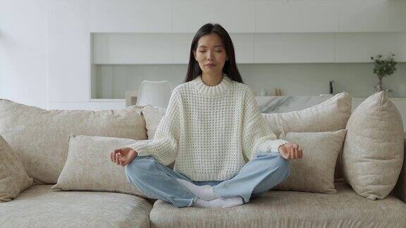 一个亚洲女孩坐在客厅的沙发上做瑜伽女瑜伽修行者莲花坐瑜伽和冥想