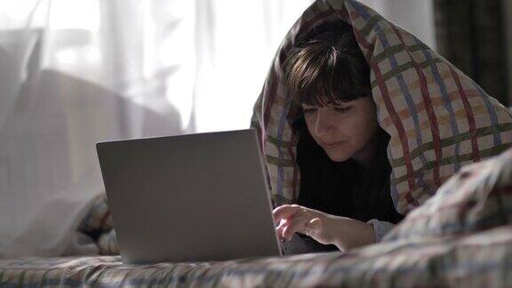 年轻女子躺在床上盖着毯子在笔记本电脑上打字