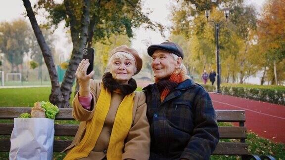 一对老夫妇爷爷奶奶坐在秋天公园的长椅上微笑着用手机自拍