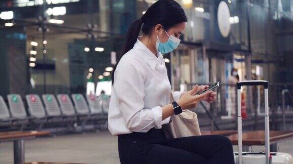 在新冠肺炎大流行期间美丽的女性戴着外科口罩坐在候机楼外的椅子上使用智能手机保持社交距离