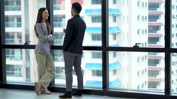 快乐的亚洲商人和女商人讨论商业战略在一个高楼大厦的现代办公室城市景观的背景两个工人在城市摩天大楼的窗前交谈