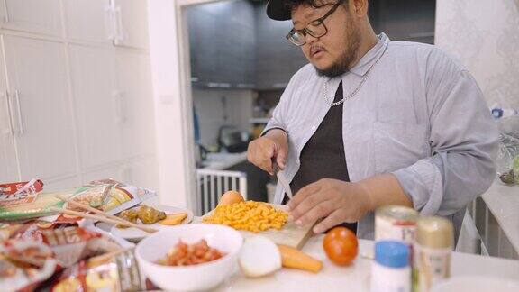 一个肥胖的男人一边欢快地唱着歌一边在厨房里把胡萝卜切成小方块放上一大堆食物准备在家做饭