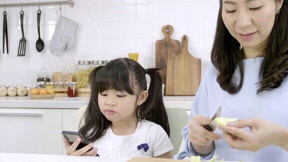 幸福的亚洲家庭妈妈正在准备蔬菜和水果女儿在家里的厨房玩智能手机健康饮食和晨间活动理念