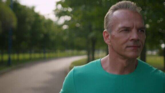 一个英俊的金发男子穿着蓝绿色的t恤手臂上挎着显示器在小公园里慢跑