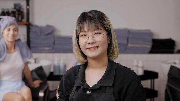 在理发店里年轻的亚洲女性发型师的肖像戴着眼镜穿着黑色的围裙面带微笑背景是美丽的亚洲客户用灰色的毛巾包着头发美容时尚个人护理事业