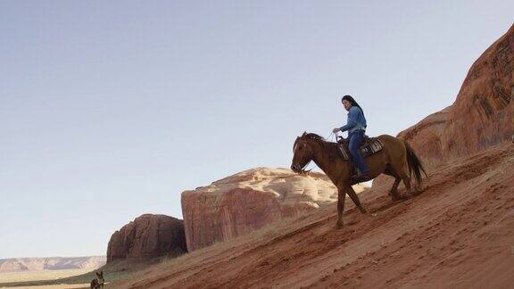 一个十几岁的美国土著女孩(纳瓦霍人)骑着她的马与她的狗在亚利桑那纪念碑谷沙漠在日落旁边的一个大的岩石形成陡峭的小山