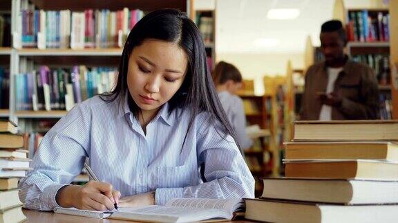 年轻美丽的亚洲女学生坐在桌子周围的书籍在大图书馆重写课本准备考试