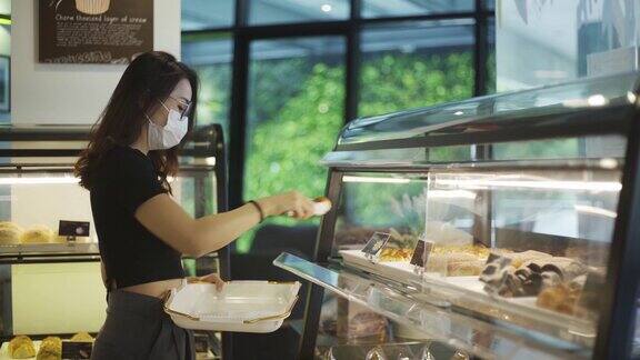 亚洲华裔美女戴着口罩在面包店挑选和购买面包糕点放在托盘上