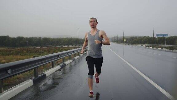 亚洲体育三项全能运动员在下雨时在高速公路上跑马拉松正面视图体格健壮的男人沿着公路跑步做有氧运动