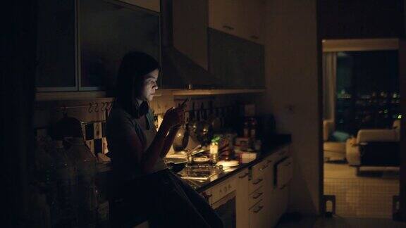 午夜时分年轻女子坐在厨房柜台上站在家里厨房里使用智能手机