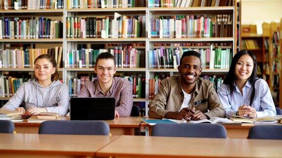 四个多民族学生的肖像坐在长桌子在大图书馆与成堆的书看着相机和正面微笑