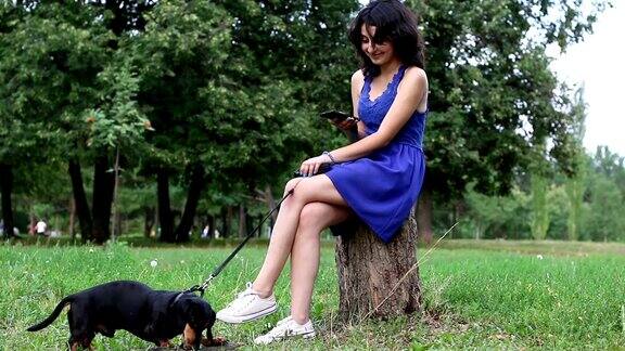 一个年轻女子坐在树桩上和一只腊肠狗在公园里用智能手机重写