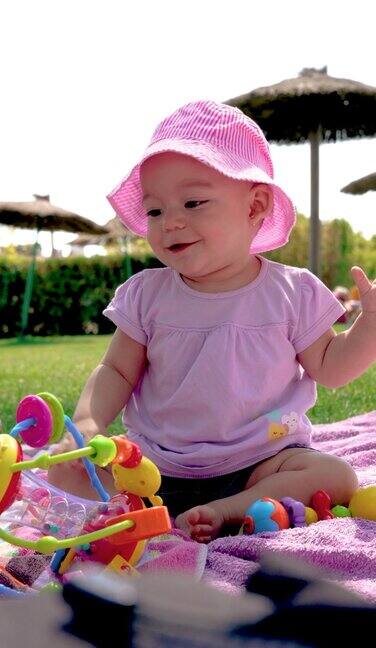 4k垂直视频中一个大眼睛的婴儿戴着一顶粉色的帽子来防晒坐在草地上的毛巾上微笑着和他的玩具玩得很开心