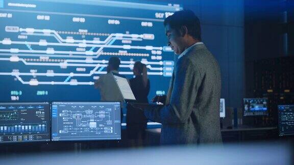英俊自信的男性项目经理站在系统控制室用笔记本电脑工作背景大屏幕显示信息图的基础设施图表系统分析侧视图弧线拍摄